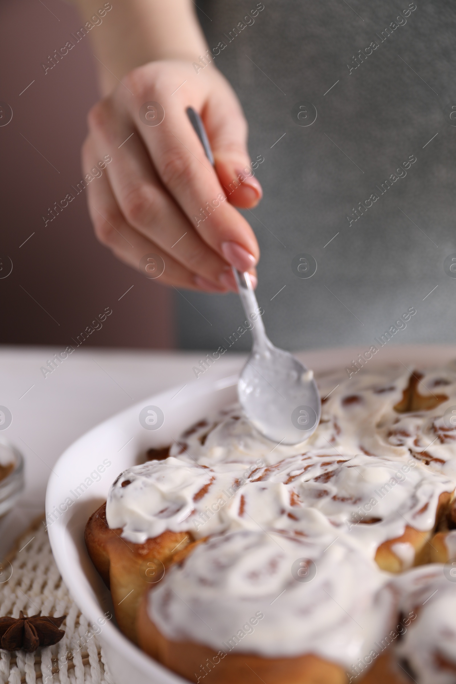 Photo of Woman spreading frosting onto freshly baked cinnamon rolls at white table against pink background, closeup