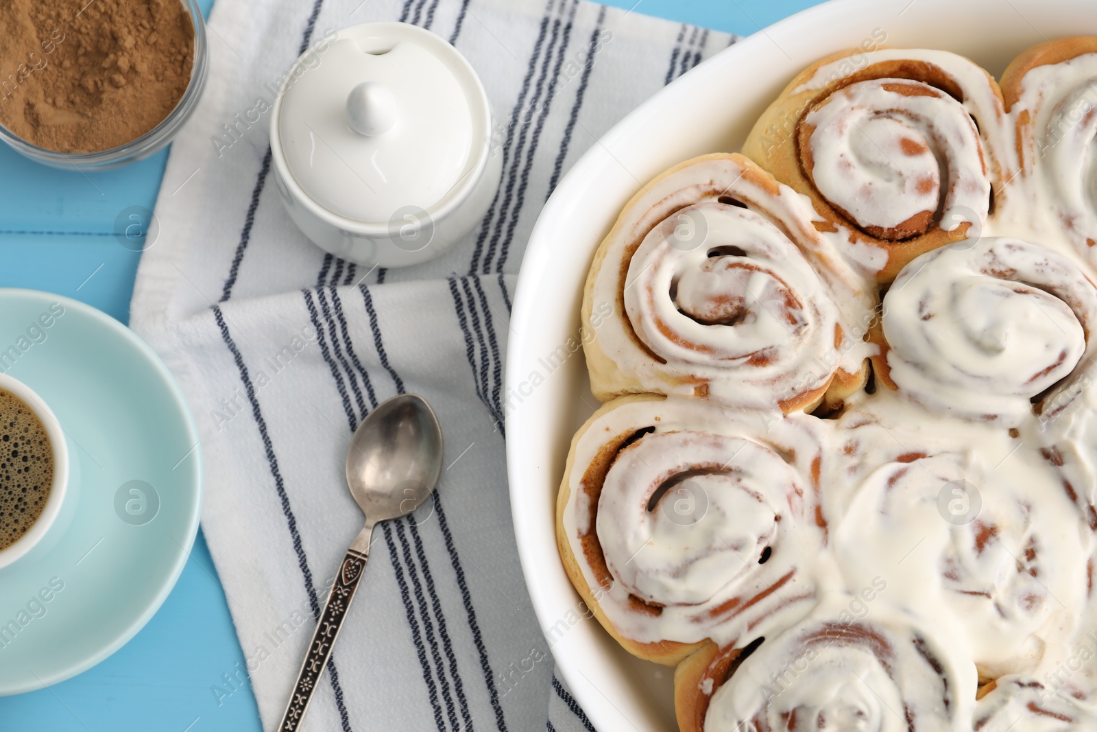 Photo of Delicious frosted cinnamon rolls in baking dish and spices on light blue wooden table, flat lay