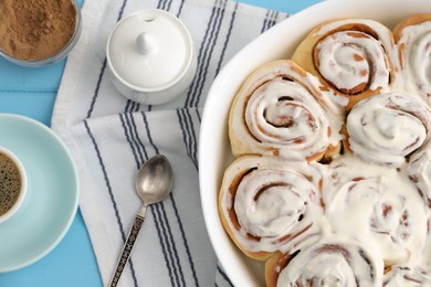 Photo of Delicious frosted cinnamon rolls in baking dish and spices on light blue wooden table, flat lay