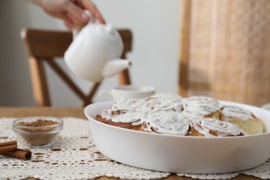 Photo of Delicious frosted cinnamon rolls and woman with teapot at table indoors, selective focus