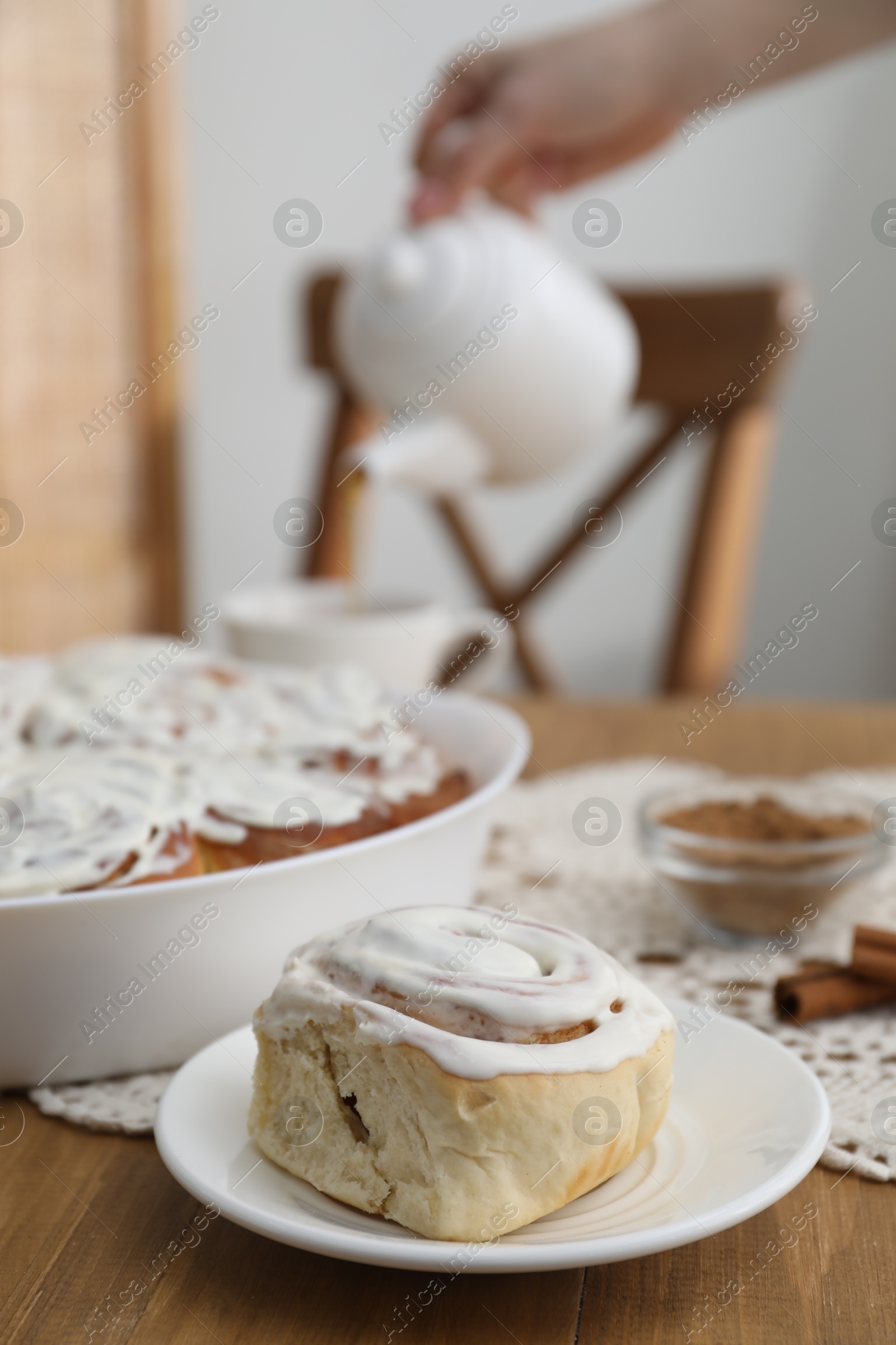 Photo of Delicious frosted cinnamon rolls and woman with teapot at wooden table indoors, selective focus