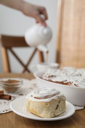 Photo of Delicious frosted cinnamon rolls and woman with teapot at wooden table indoors, selective focus