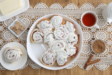 Photo of Delicious frosted cinnamon rolls, spices, tea and butter on wooden table, flat lay