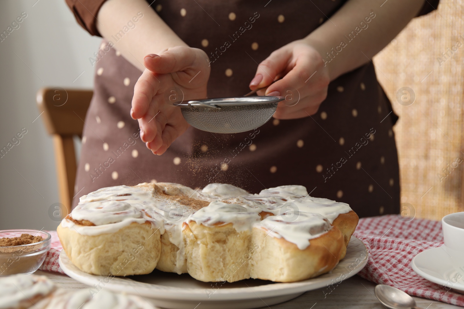 Photo of Woman with sieve adding cinnamon powder onto freshly baked rolls at table indoors, closeup