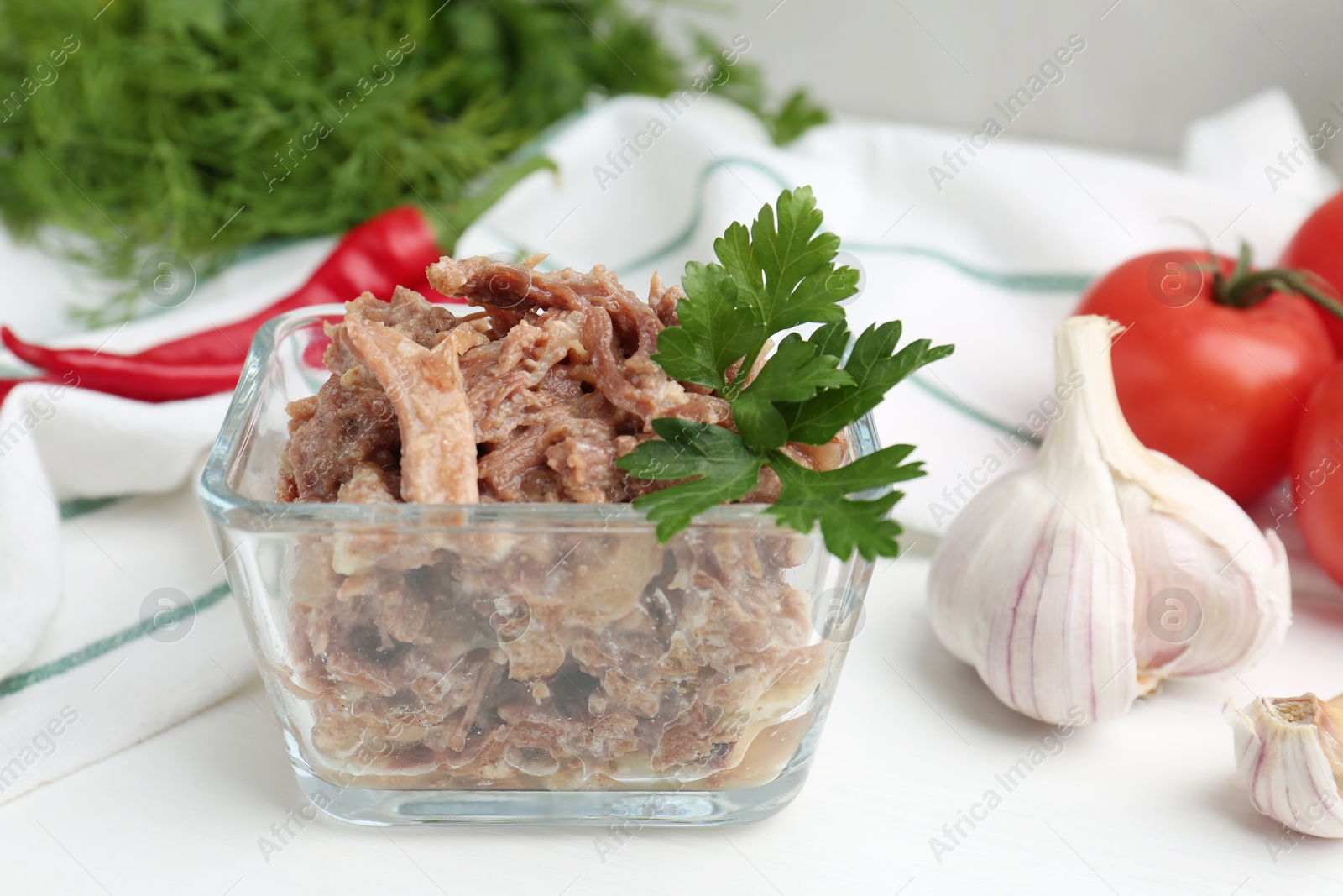 Photo of Canned meat with parsley in bowl and fresh vegetables on white wooden table, closeup