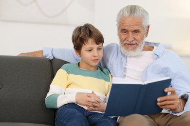 Photo of Grandpa and his grandson reading book together on sofa at home