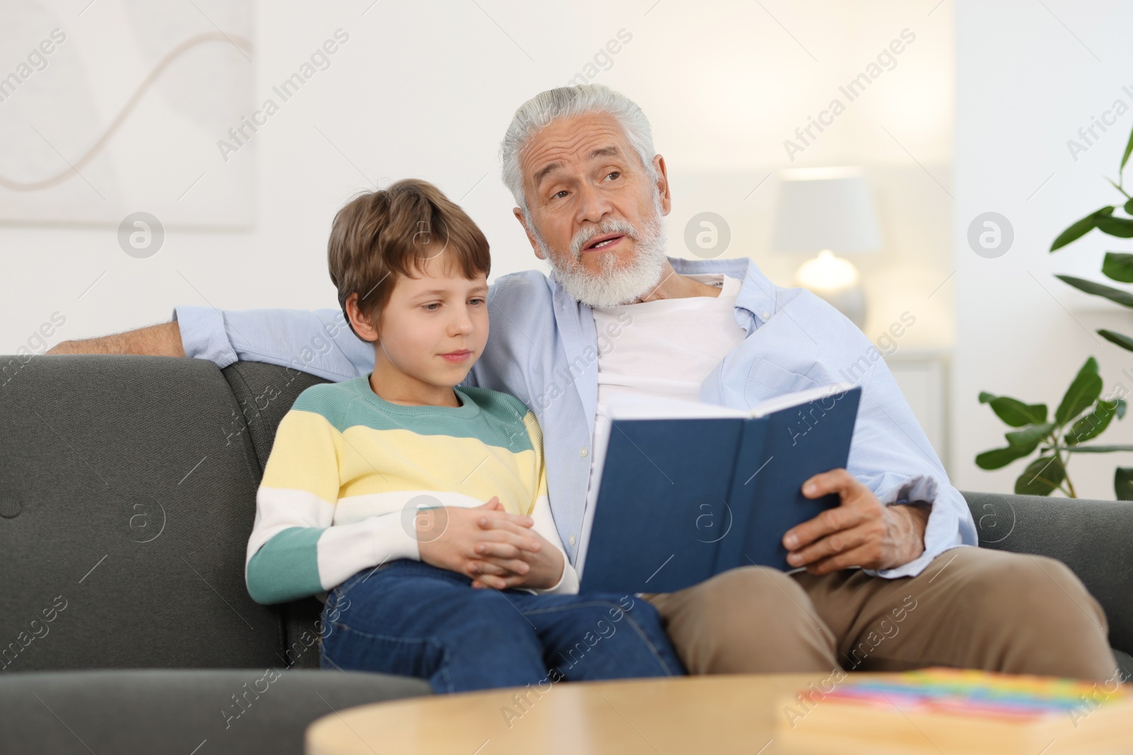 Photo of Grandpa and his grandson reading book together on sofa at home