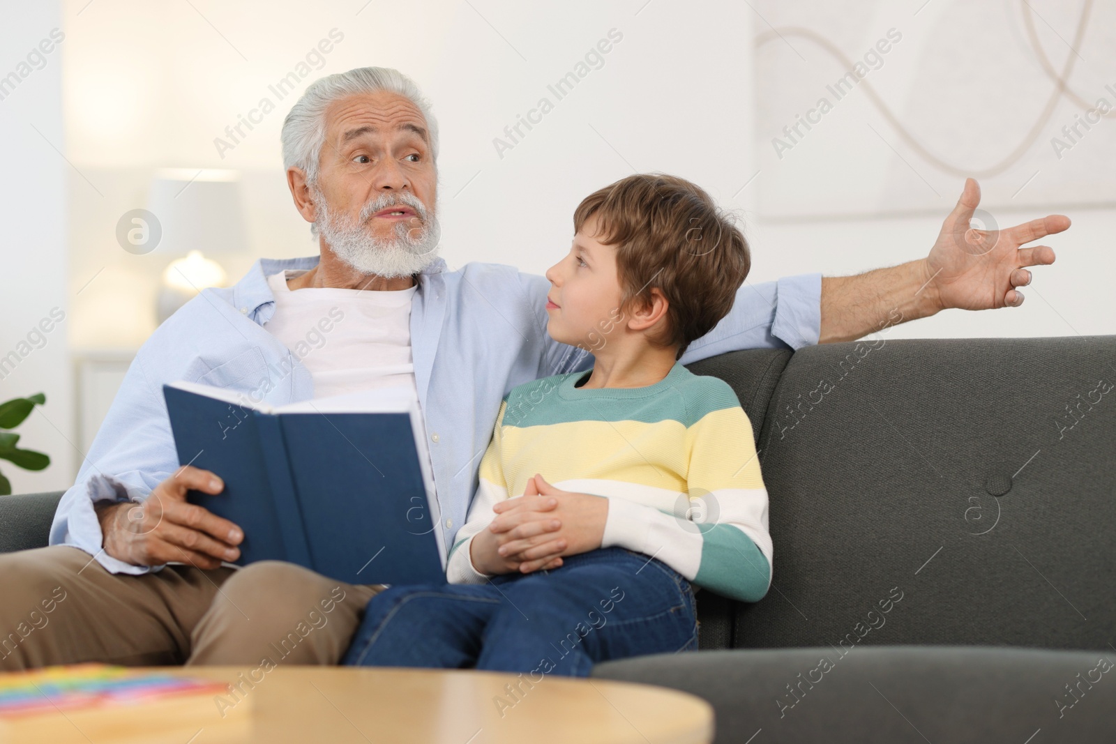 Photo of Grandpa and his grandson reading book together on sofa at home