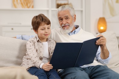 Photo of Grandpa and his grandson reading book together on sofa at home