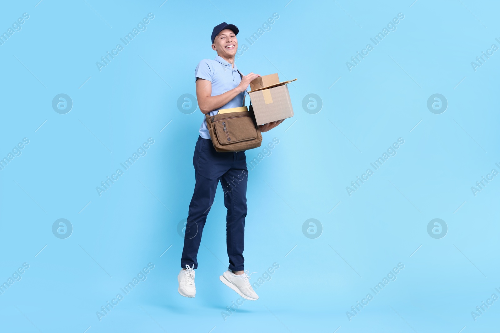 Photo of Happy postman jumping with bag and parcels on light blue background