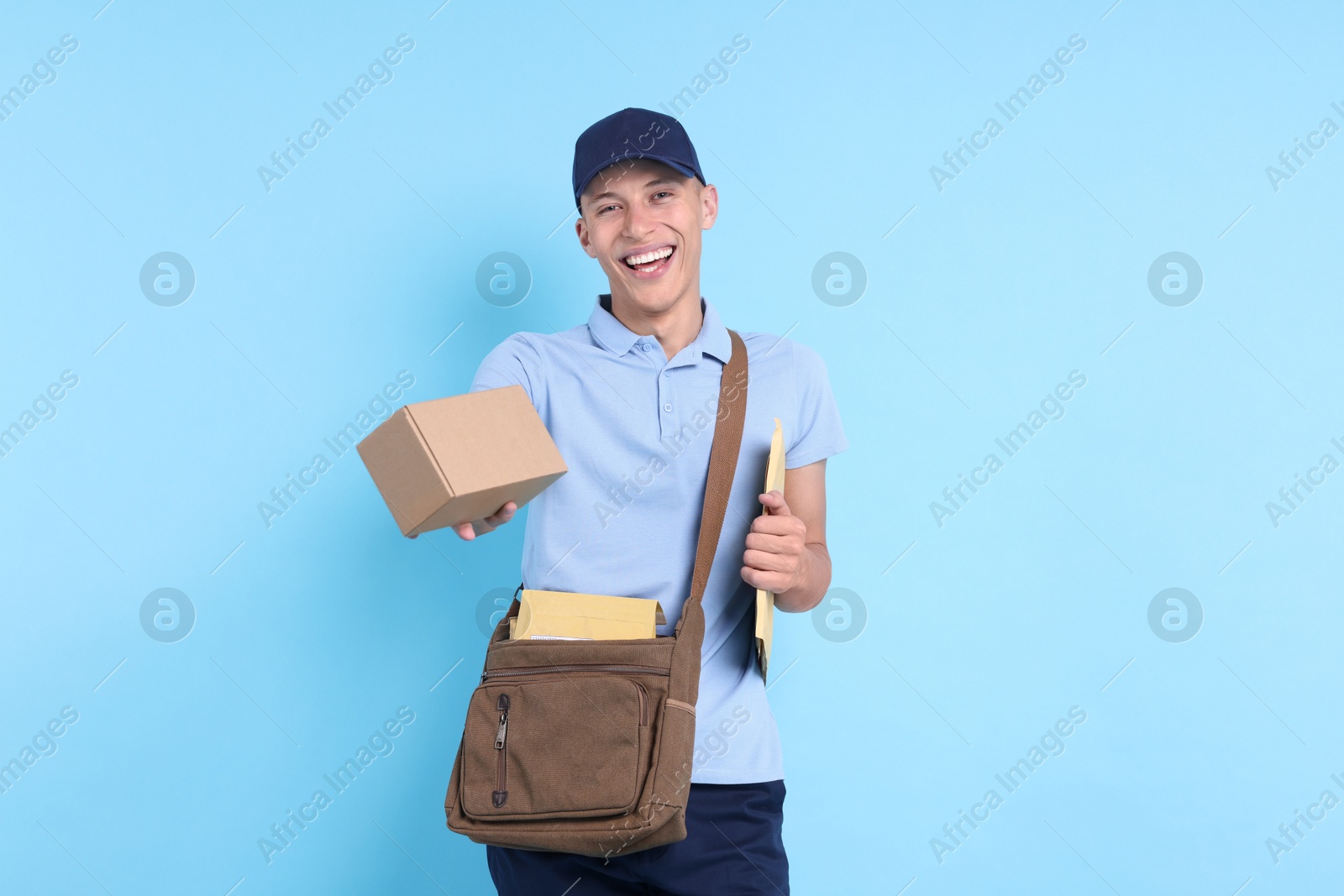 Photo of Happy postman with bag, envelopes and parcel on light blue background