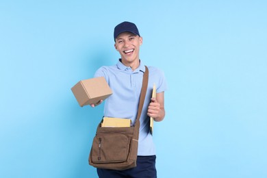 Happy postman with bag, envelopes and parcel on light blue background