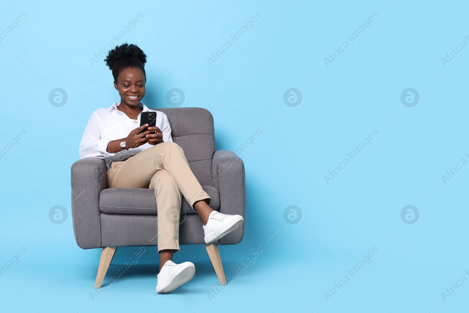 Photo of Smiling woman with smartphone sitting in armchair on light blue background. Space for text