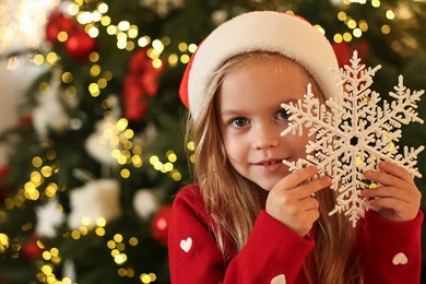 Little girl in Santa hat with decorative snowflake at home, space for text. Christmas celebration