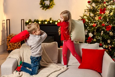 Photo of Little kids having pillow fight on sofa in room decorated for Christmas