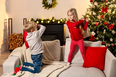 Little kids having pillow fight on sofa in room decorated for Christmas