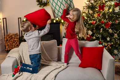 Little kids having pillow fight on sofa in room decorated for Christmas