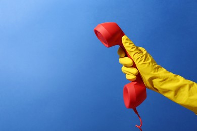 Woman in rubber glove holding red telephone handset on blue background, closeup. Space for text