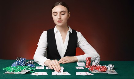 Professional croupier with casino chips and playing cards at gambling table on color background