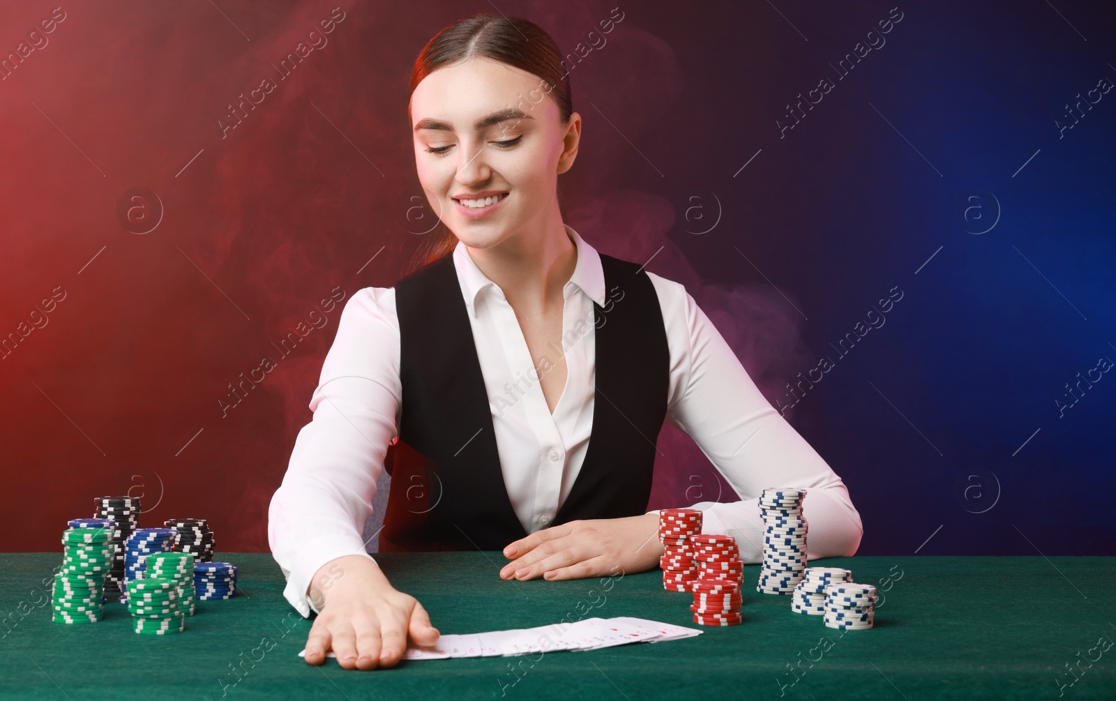 Photo of Professional croupier with casino chips and playing cards at gambling table on color background with smoke