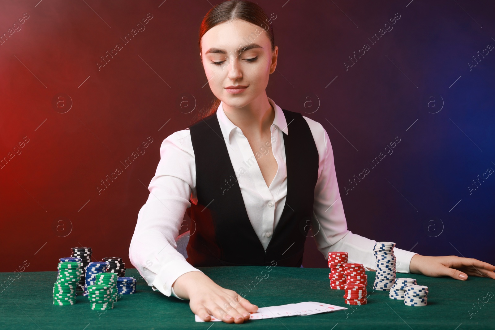 Photo of Professional croupier with casino chips and playing cards at gambling table on color background with smoke