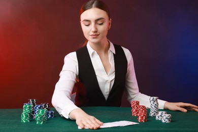 Professional croupier with casino chips and playing cards at gambling table on color background with smoke