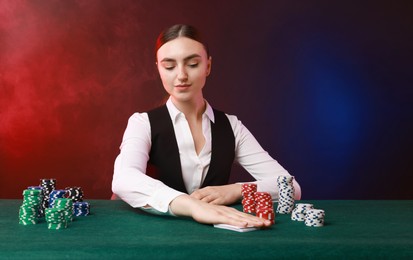 Professional croupier with casino chips and playing cards at gambling table on color background with smoke