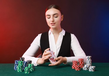 Professional croupier with chips shuffling playing cards at gambling table on color background