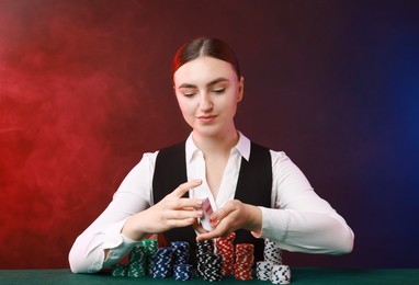 Photo of Professional croupier with chips shuffling playing cards at gambling table on color background with smoke