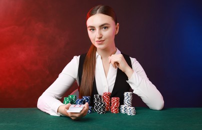 Photo of Professional croupier with casino chips and playing cards at gambling table on color background with smoke