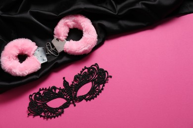 Photo of Fluffy handcuffs, lace mask and black fabric on pink background, flat lay. Space for text