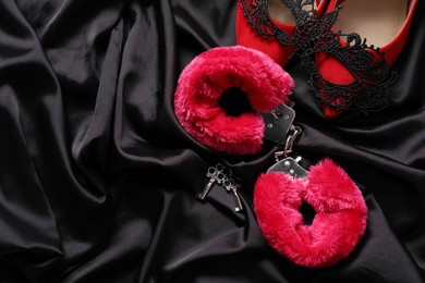 Photo of Red fluffy handcuffs, keys, shoes and lace mask on black fabric, flat lay. Space for text