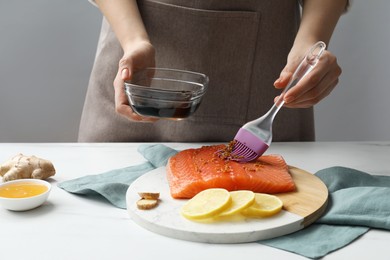 Photo of Woman marinating salmon fillet in dish at white table, closeup