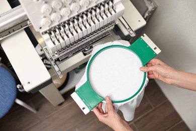 Photo of Woman using embroidery machine to make design on T-shirt indoors, closeup