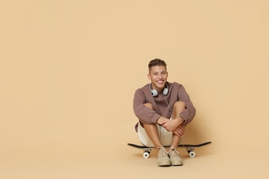 Photo of Happy man with headphones sitting on skateboard against beige background. Space for text