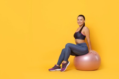 Photo of Smiling woman in sportswear sitting on fitness ball against yellow background. Space for text