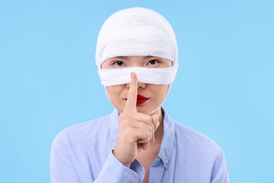 Woman with nose wrapped in medical bandage after plastic surgery operation making silent gesture on light blue background