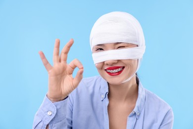 Woman with nose wrapped in medical bandage after plastic surgery operation showing ok gesture on light blue background