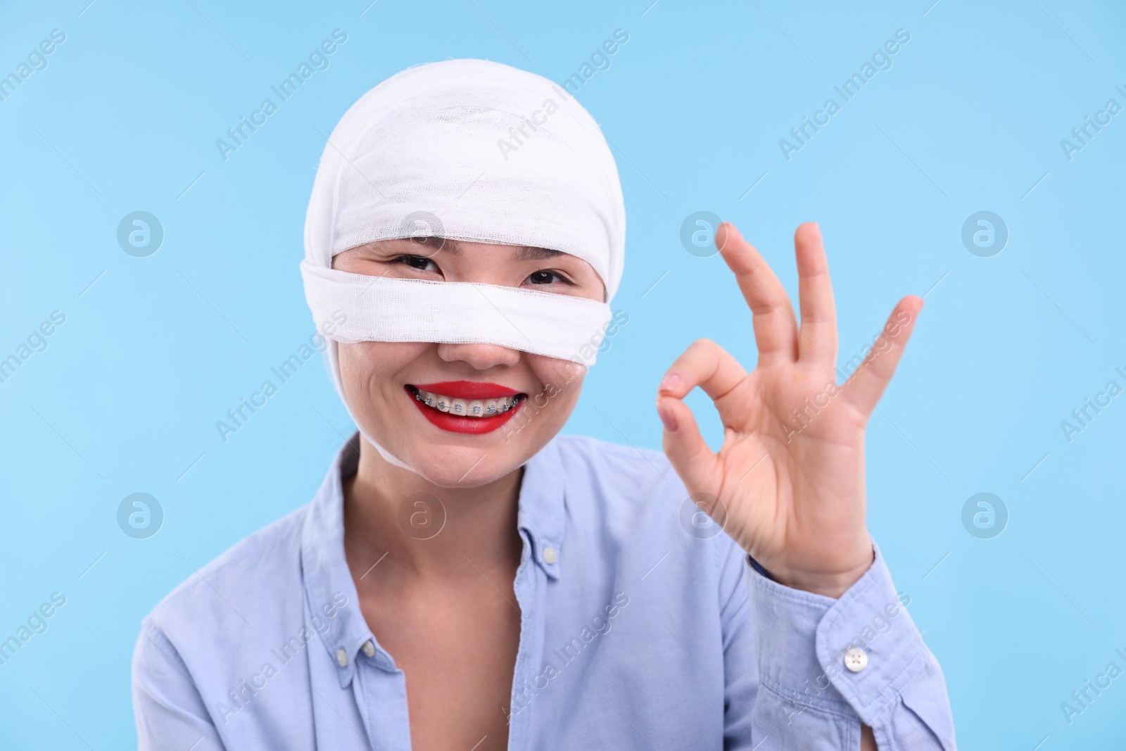 Photo of Woman with nose wrapped in medical bandage after plastic surgery operation showing ok gesture on light blue background