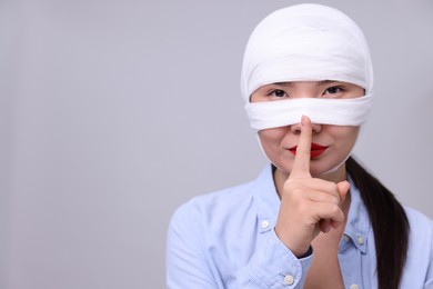 Photo of Woman with nose wrapped in medical bandage after plastic surgery operation making silent gesture on light grey background. Space for text