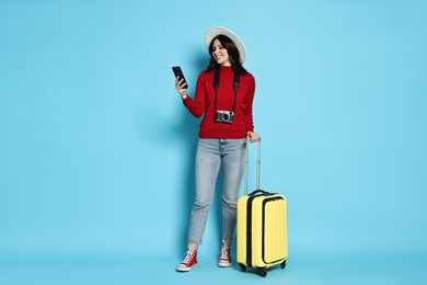 Young tourist in hat with camera, phone and suitcase on light blue background