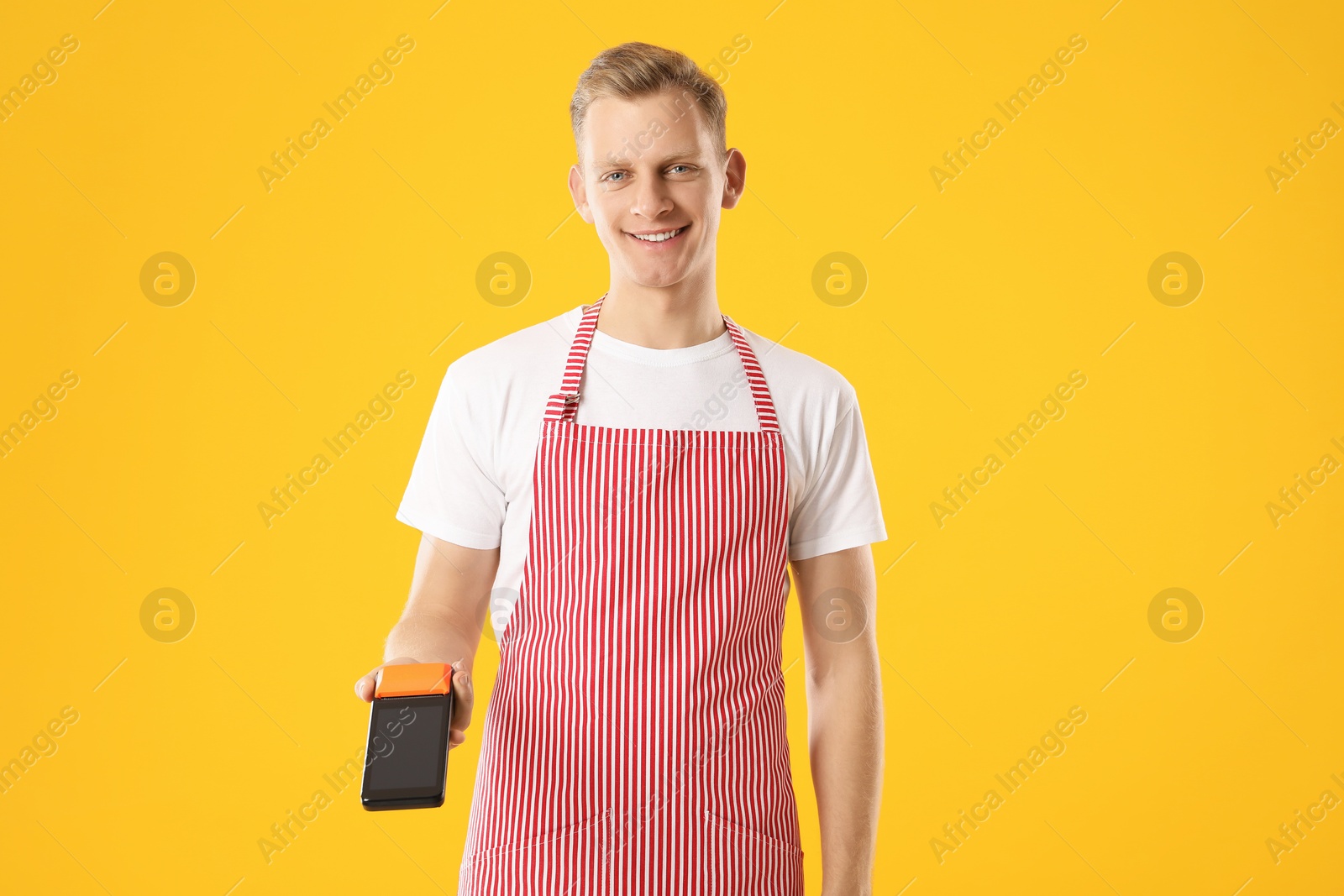 Photo of Happy young man in apron with payment terminal on yellow background