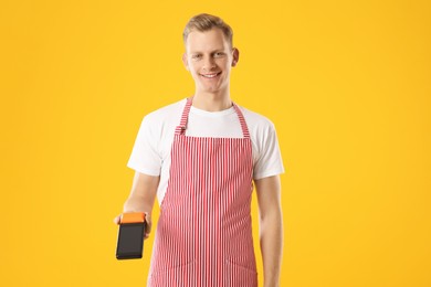 Photo of Happy young man in apron with payment terminal on yellow background