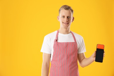 Photo of Happy young man in apron with payment terminal on yellow background, space for text