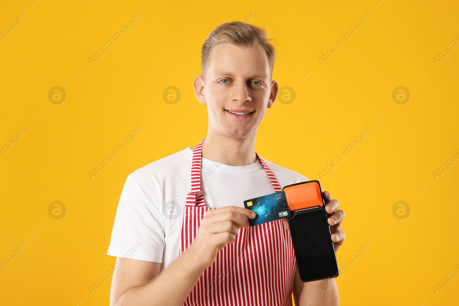 Photo of Happy young man in apron with payment terminal and debit card on yellow background