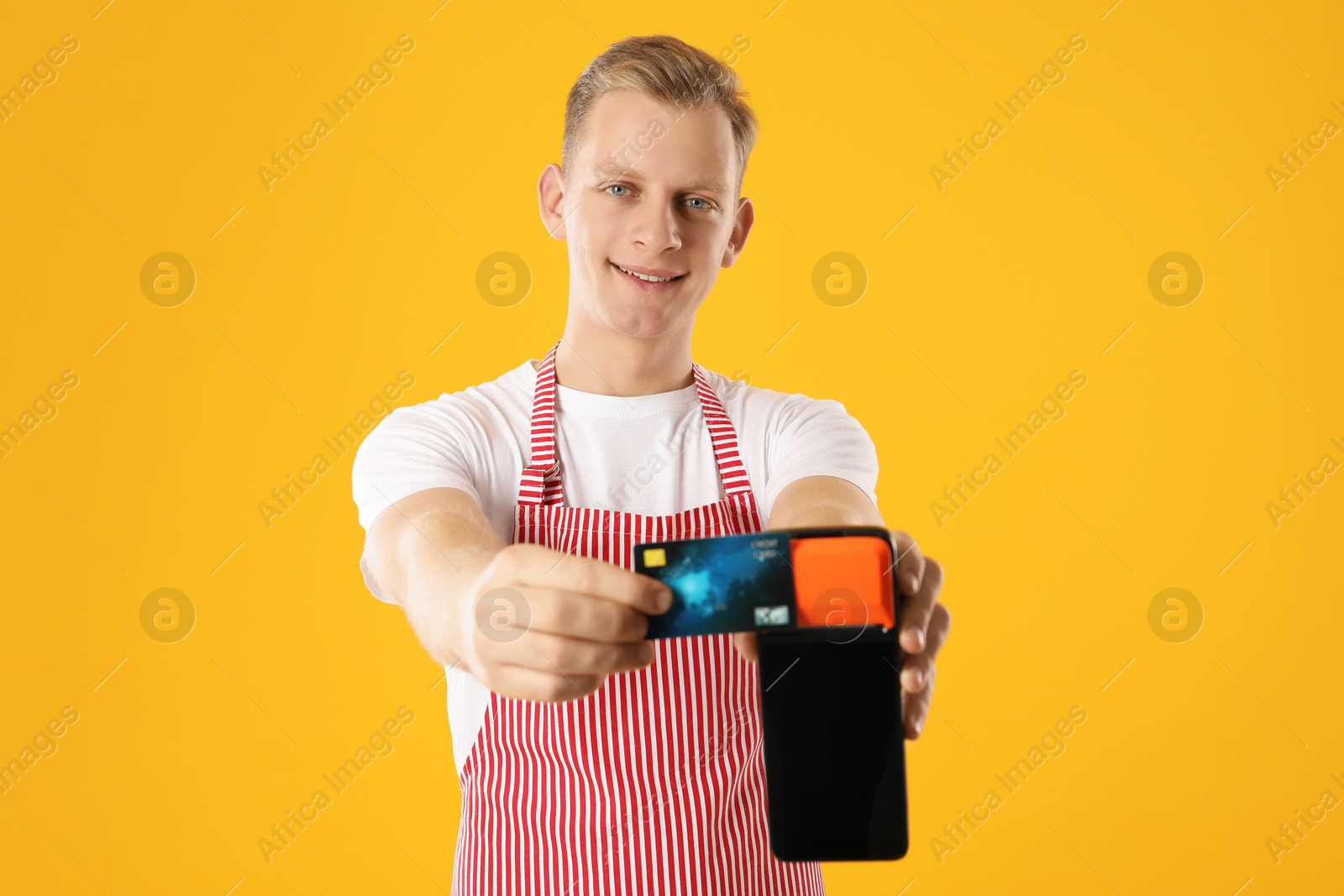 Photo of Happy young man in apron with payment terminal and debit card on yellow background