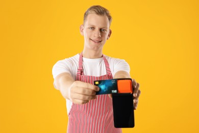 Photo of Happy young man in apron with payment terminal and debit card on yellow background