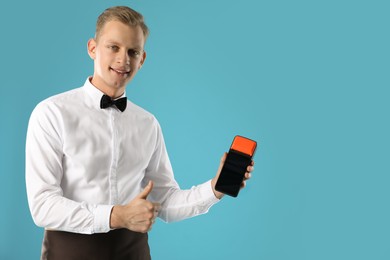 Photo of Happy waiter with payment terminal showing thumbs up on light blue background, space for text