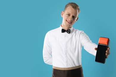 Happy waiter with payment terminal on light blue background, space for text