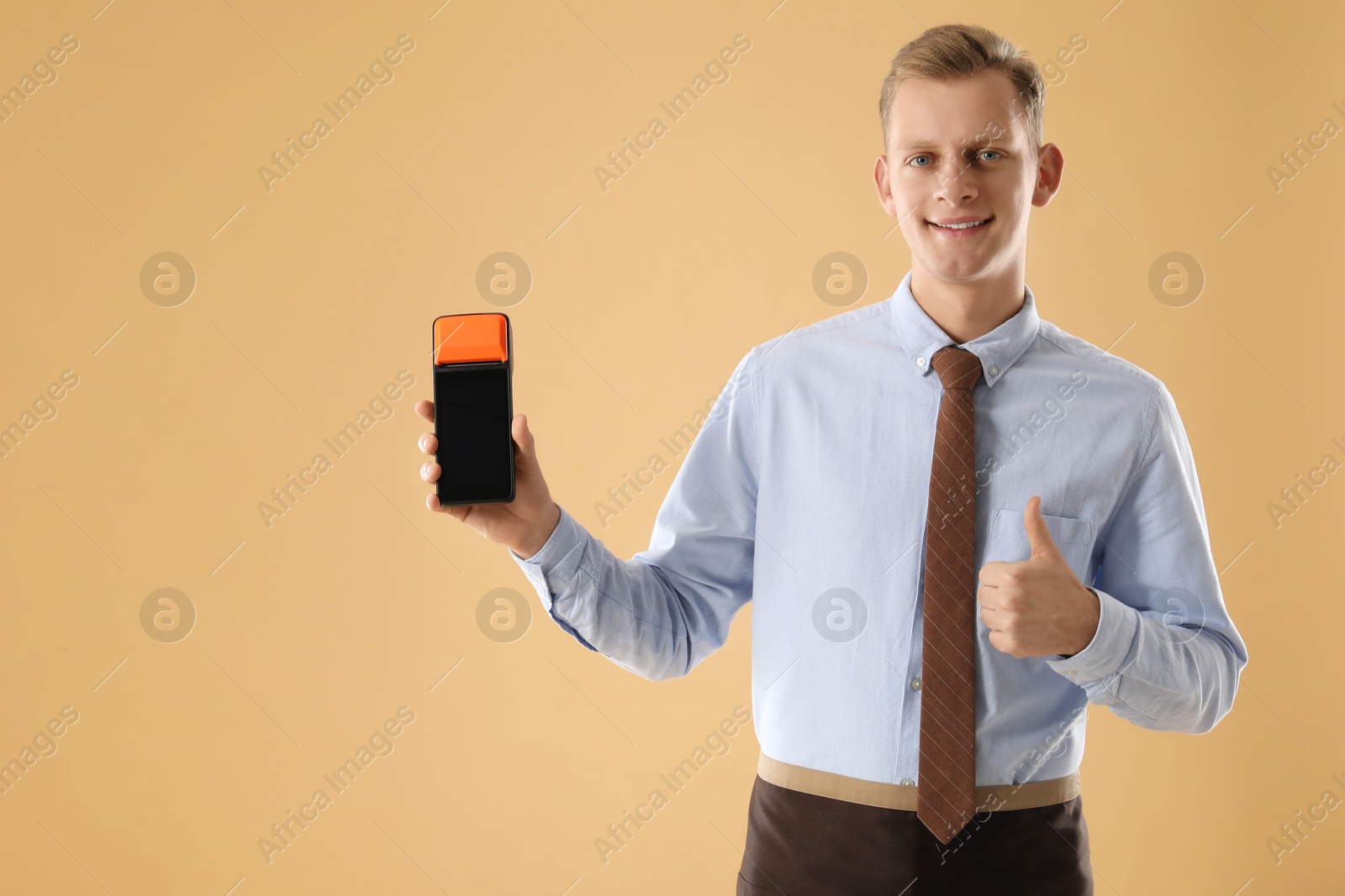 Photo of Happy young man with payment terminal showing thumbs up on beige background, space for text